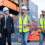 Logistics consulting team walking through a shipping yard, discussing operations with professionals in safety gear.