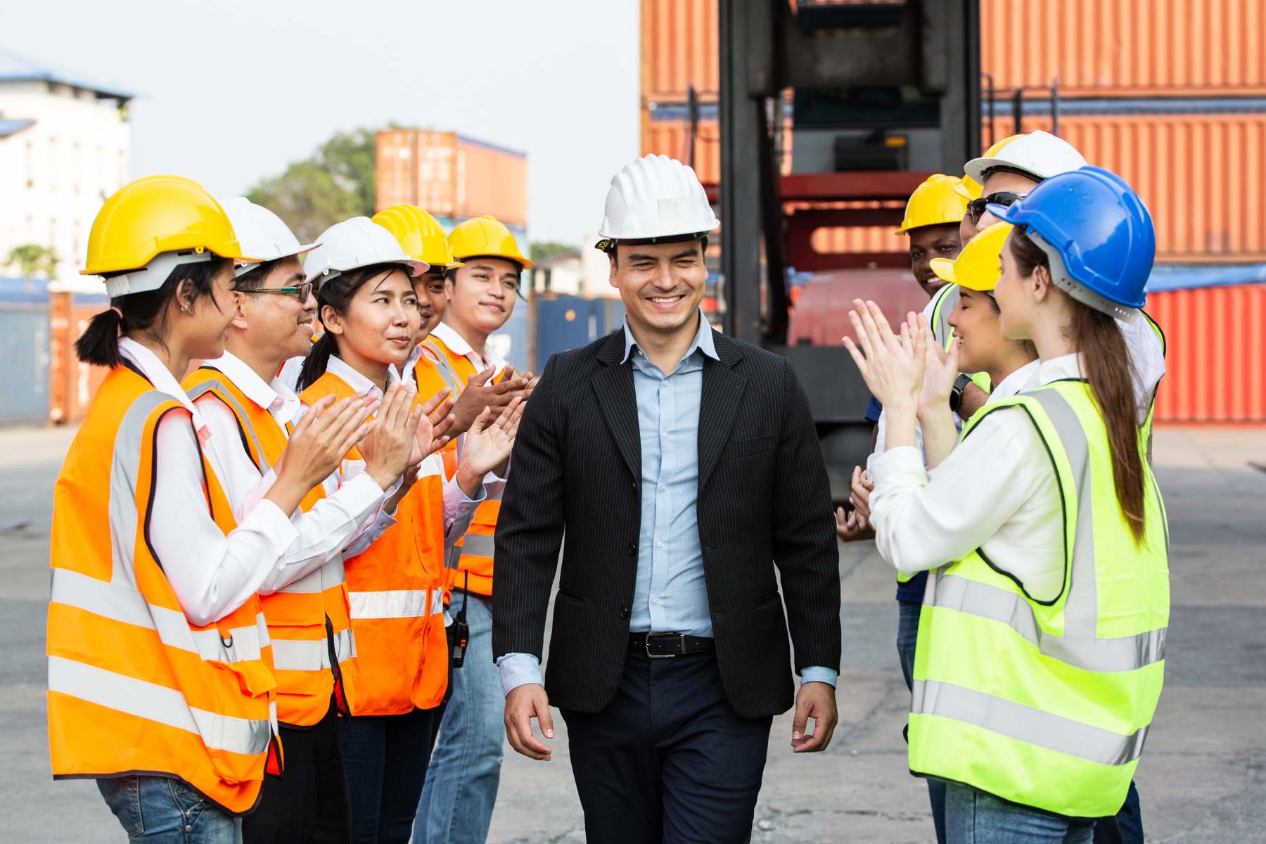 Team of logistics professionals applauding and welcoming a leader in a warehouse environment with shipping containers in the background.